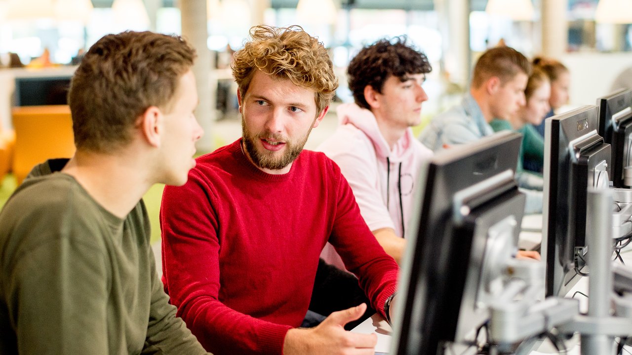 Twee studenten aan het praten achter computer in mediatheek